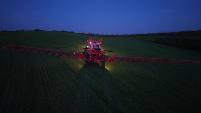 Rear aerial view of a crop sprayer moving away across a field at blue hour, with broad red booms spanning the planted rows and tractor lights shining ahead.