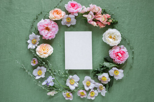Overhead view of blank card inside a floral wreath of pink roses, anemones, and foliage on a green tablecloth