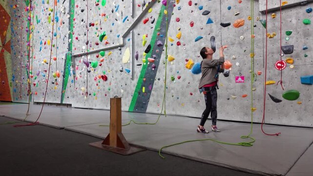 Female rock climber ascending an indoor climbing wall with colorful holds, wearing a safety harness and climbing shoes in a modern climbing gym.