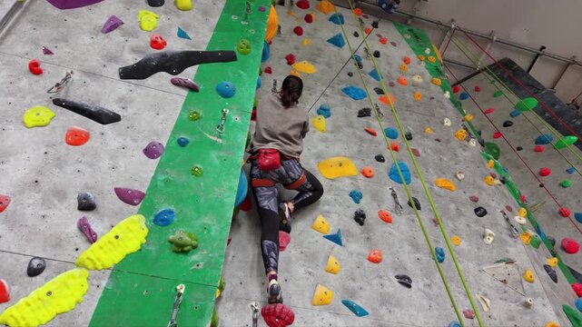 Female rock climber ascending an indoor climbing wall with colorful holds, wearing a safety harness and climbing shoes in a modern climbing gym.