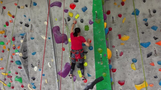 Female rock climber ascending an indoor climbing wall with colorful holds, wearing a safety harness and climbing shoes in a modern climbing gym.