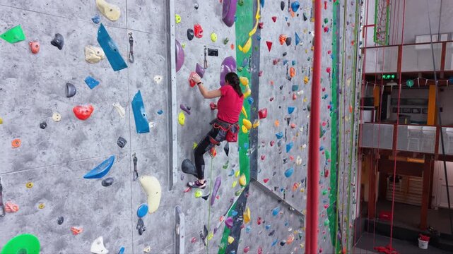 Female rock climber ascending an indoor climbing wall with colorful holds, wearing a safety harness and climbing shoes in a modern climbing gym.