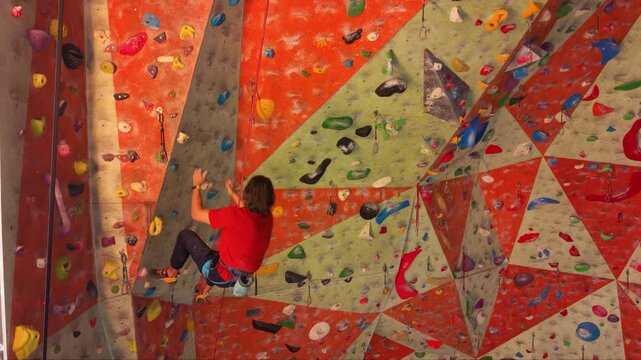Indoor rock climber ascending a steep artificial climbing wall with colorful holds and volumes while secured by a rope and harness, showing strength, technique and concentration during sport climbing.