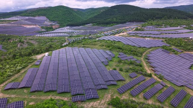 Aerial view of a vast solar power plant covering rolling hills and valleys with thousands of photovoltaic panels arranged in long rows beside a winding road and green mountain landscape.