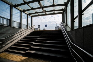 Fototapeta premium Concrete stairs lead from underground subway exit to city street level. Glass roof covers pedestrian passage staircase with metal railings. Sunny day view includes outdoor crosswalk sign, modern