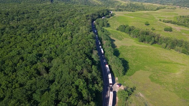 Aerial view of a long traffic queue with trucks and cars moving slowly along a winding two-lane road through dense green forest and rolling countryside, showing heavy congestion on a rural route.