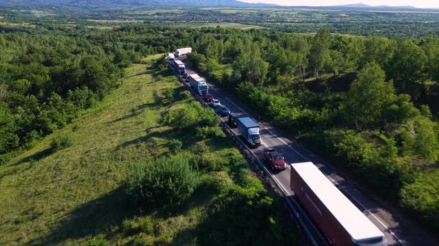 Aerial view of a long traffic queue with cars and heavy trucks on a winding rural mountain road, surrounded by dense green forest and countryside.
