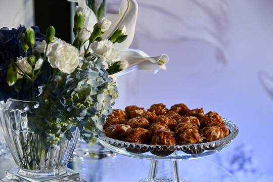 gourmet caramelized nut sweets on crystal glass stand next to white Lilium lilies Dianthus caryophyllus and blue Hydrangea macrophylla flowers close up, luxury party dessert table setup stock photogra