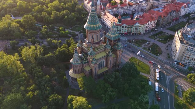 Aerial golden hour views of the Metropolitan Cathedral in Timișoara, Romania, showing its distinctive patterned roof, tall central tower and surrounding green parkland, roads and historic city neighbo