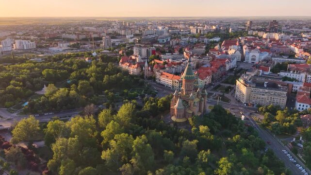Aerial view of the Romanian Orthodox Metropolitan Cathedral in Timișoara, Romania, surrounded by dense green parkland, city streets and historic buildings in warm golden-hour light.
