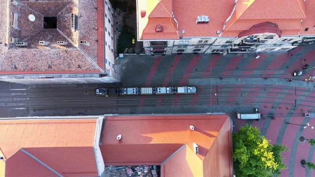 Top-down aerial view of a modern tram travelling through the historic city centre of Timișoara, Romania, crossing a pedestrian square surrounded by traditional European architecture.