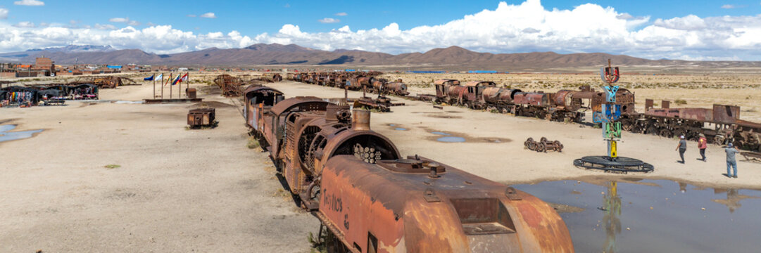 Train Cemetery featuring rusted locomotive skeletons and abandoned railway carriages scattered across the arid desert landscape. Aerial view.