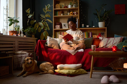 Young adult African American woman sitting on sofa and reading book with dog lying on floor. Image represents mental wellbeing and leisure, suitable for lifestyle marketing campaigns