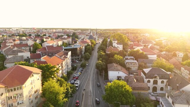 Aerial view of an urban boulevard with public buses, tram tracks and historic churches surrounded by residential buildings and trees in warm sunset light.