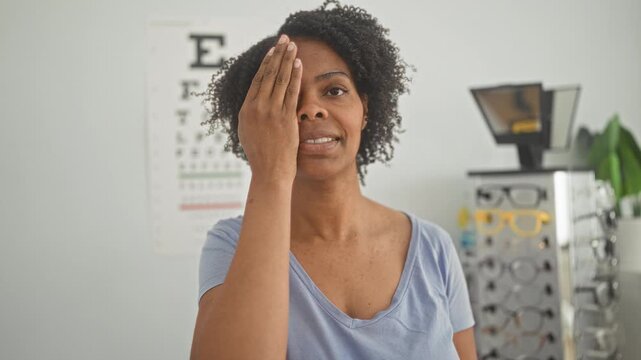 Woman covers one eye with hand for vision test in building, standing before eye chart and glasses display; calm checkup.