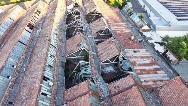 Aerial view of a severely damaged and partially collapsed industrial building roof, showing broken structural frames, deteriorated tiles and metal roofing on an abandoned factory complex.