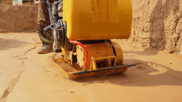Construction worker operating a yellow vibratory plate compactor, leveling and preparing the ground for a new building foundation, ensuring stable soil compaction on a construction site