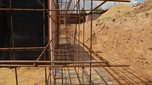 Construction site with an excavated trench, showing rebar cages prepared for pouring concrete foundation, alongside a foundation wall covered with black waterproofing membrane