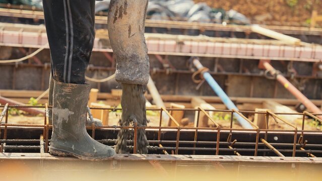Worker wearing protective rubber boots standing on formwork while pouring fresh concrete mix from a pump hose into a building foundation structure with rebar