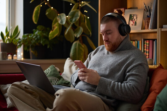 Young adult Caucasian man wearing headphones using smartphone while sitting on sofa with laptop. Image represents remote work and digital lifestyle, suitable for technology marketing