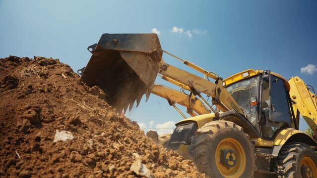 Yellow backhoe loader moving and scooping brown dirt, performing groundworks at a building area against a clear blue sky, demonstrating heavy equipment at work