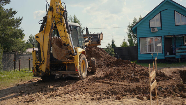 Yellow excavator actively digging and moving soil on a residential construction site, preparing the ground next to a blue house under a bright sky