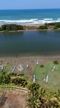Aerial view of traditional fishing boats moored on the sandy shore near a river estuary, surrounded by lush palm trees and coastal vegetation in Kecamatan Kebumen, Jawa Tengah, Indonesia.