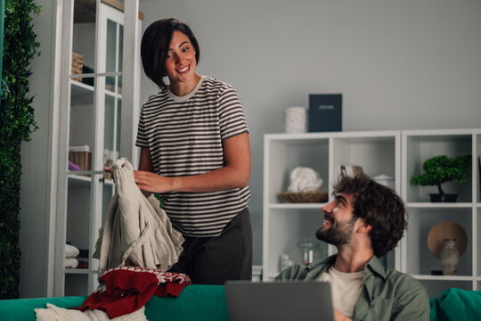Happy couple interacting at home, woman folding laundry while man works on laptop, sharing a domestic moment