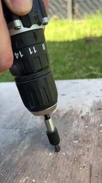 Close up view of a worker's hand using a cordless electric screwdriver to drive a screw into a wooden plank during an outdoor repair project on a grassy background.