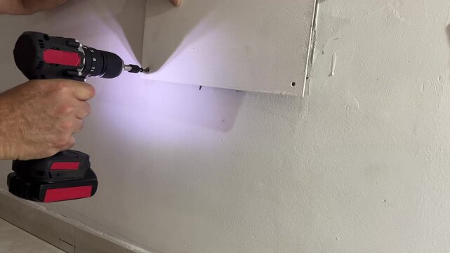 Handyman using a cordless electric screwdriver to mount a plasterboard panel on the wall with the tool's led light illuminating the screw