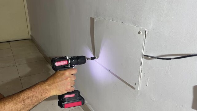 Skilled handyman's hands using a professional cordless power screwdriver with a flashlight to install a board indoors doring home renovation