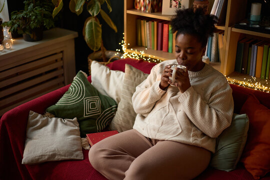 Young adult African American woman sitting on sofa drinking hot beverage, enjoying quiet evening at home. Image represents mental wellbeing and self care marketing campaigns