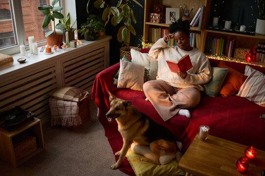 Young adult African American woman sitting on sofa reading book with dog nearby, representing mental wellbeing and leisure. Image suitable for lifestyle blogs and pet care marketing