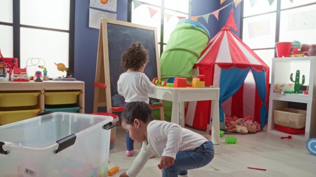 Little boy picking up plastic blocks by a toy tent and chalkboard in a preschool classroom; playful learning curiosity.