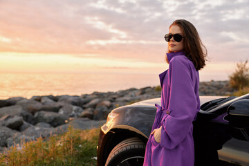 Fototapeta premium Woman lifestyle scene at seaside during sunset, wearing a purple coat and sunglasses, leaning beside a sleek car, captures confident pose and relaxed mood in golden hour light