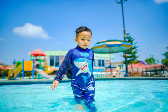 A cheerful Asian boy in a blue shark swimsuit smiles while playing in a sunny outdoor swimming pool at a resort.
