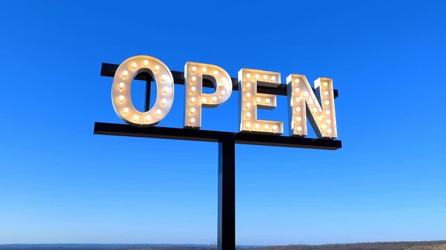 Illuminated OPEN marquee sign mounted on black metal pole against clear blue sky, warm bulb glow, bold lettering, and simple roadside storefront display with wide empty space