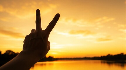 Fototapeta premium Hand forms V sign against warm golden sunset sky over calm water. Gesture symbolizes peace victory hope success and togetherness. Positive outdoor moment.