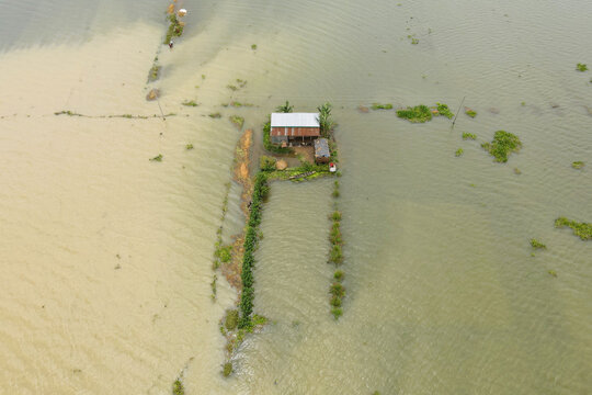 Aerial view of a house isolated by floodwaters with contrasting muddy and clear water, green vegetation, and a small boat in Sunamganj, Sylhet Division, Bangladesh.