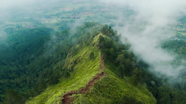 Aerial view of the lush green ridge of Gunung Andong with a narrow hiking trail surrounded by misty clouds and dense forest in Kecamatan Ngablak, Jawa Tengah, Indonesia.