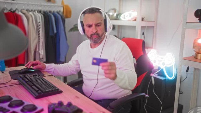 Man holding creditcard and typing on keyboard while wearing headset at gaming desk with neon controller sign in studio; focused online purchase.