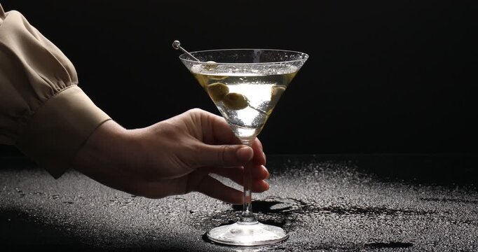 Woman putting skewer with fresh olives into glass with chilled martini cocktail and taking it from wet mirror table against black background, closeup