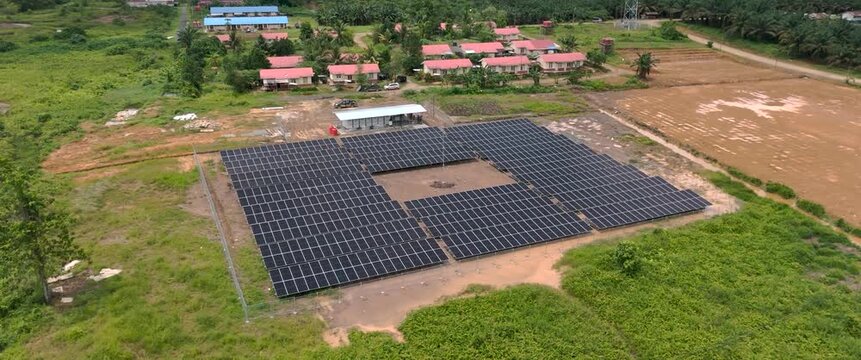 Aerial view of a solar power plant with rows of photovoltaic panels situated in a rural landscape near residential buildings in Tanahgrogot, Kalimantan Timur, Indonesia.