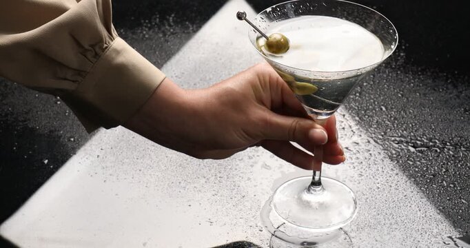 Woman putting skewer with fresh olives into glass with chilled martini cocktail and taking it from wet mirror table, closeup