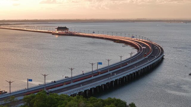 Aerial view of the curved Mandara Toll Road over the water at sunset in Kabupaten Badung, Bali, Indonesia.
