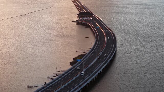 Aerial view of the Bali Mandara Toll Road crossing the water with the Nusa Dua Toll Plaza at sunset in Kabupaten Badung, Bali, Indonesia.
