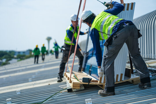 A solar installation engineer in a white helmet and high visibility vest carefully bends over the pallet checking each module label on the curved metal roof of an industrial factory.