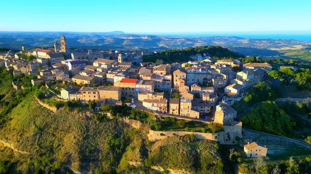 Stupendous aerial push-in pedestal down shot from Monterubbiano, approaching the town centre with its vintage Marche buildings, churches, belltowers, clocktowers, in the afternoon sunlight, with trees