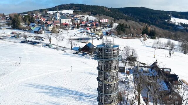 Aerial view of the snow-covered Majak Jary Cimrmana observation tower surrounded by winter forests and hills in Korenov, Liberecky kraj, Czechia.