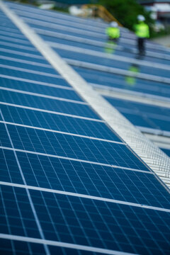 Two renewable energy specialists in hard hats and hi vis workwear quietly move small and distant across the vast glowing array high on an industrial warehouse roof in a Thai city.